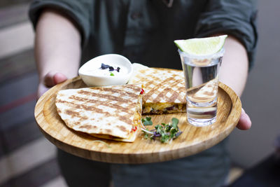 The glass of tequila with lime and portion of quesadilla on round wooden board in hands