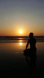 Silhouette man sitting on beach against sky during sunset