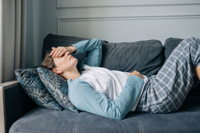 A stressed woman with a migraine lies on the sofa at home, covering her face with her hand.