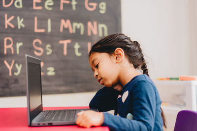 Side view of diverse pre school age girl at home doing distance learning on laptop computer