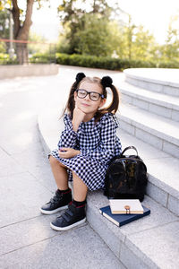 Smiling happy kid girl 5-6 year old wear uniform dress and glasses sitting on stair with books
