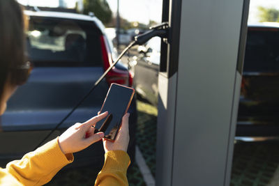 Woman using smart phone at electric vehicle charging station on sunny day