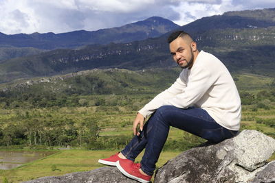 Young man sitting on landscape against mountains