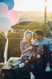 Couple sitting with wine by balloons against sky