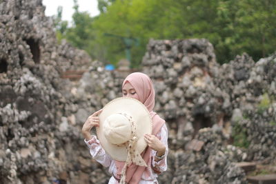 Portrait of muslim woman wearing hijab holding hat standing against plants