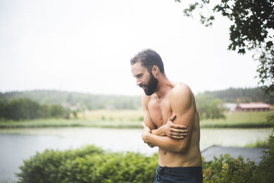 Young man looking away while standing by lake against trees