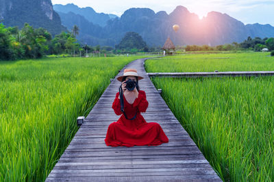 Asian woman holding a camera sits on a wooden walkway with green rice fields in vang vieng, laos