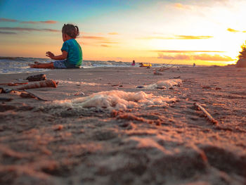 Little girl sitting on shore at beach against sky during sunset