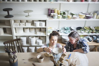 High angle view of mature female potter looking at young craftsperson molding clay at workshop