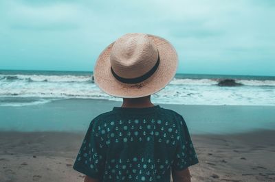 Rear view of boy wearing hat while standing at beach against sky