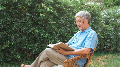 Side view of man reading book while sitting on chair against plants