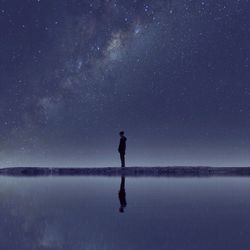 Reflection of silhouette man standing by lake against stars in sky on water