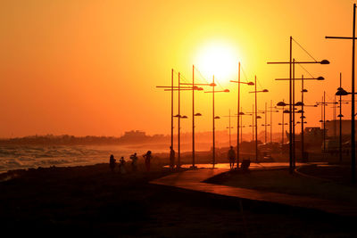 Silhouette people by street lights at beach against orange sky