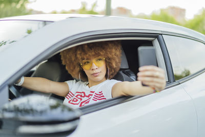 Woman with afro hair taking a photo with her smartphone next to her car
