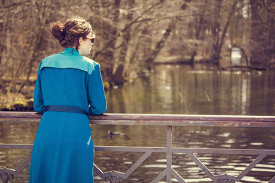 Rear view of woman in blue trench coat standing by lake during sunny day