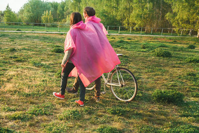 Young couple in pink raincoat walking with bicycle on field against sky