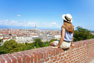 Beautiful young woman with hat sitting on wall looking at turin cityscape, italy