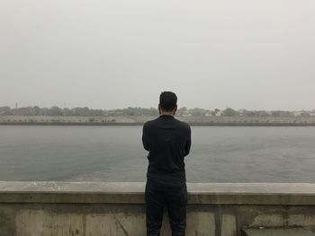 Rear view of man standing at retaining wall by river against clear sky