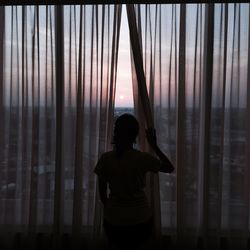 Rear view of woman looking at cityscape through curtain