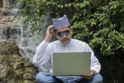 Mature man wearing sunglasses while using laptop against waterfall