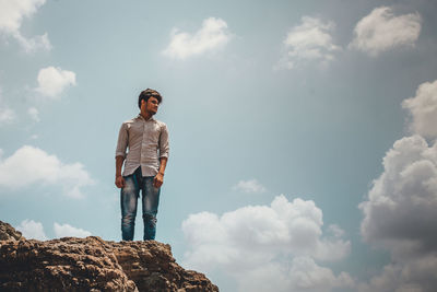 Low angle view of young man standing on rock against sky