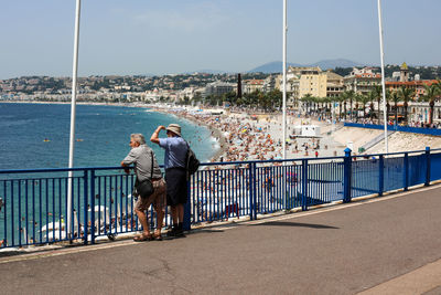 People on railing by sea against sky in city