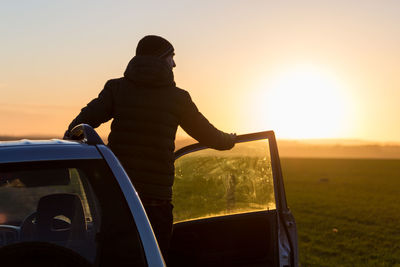 Rear view of man standing on car against sky during sunset