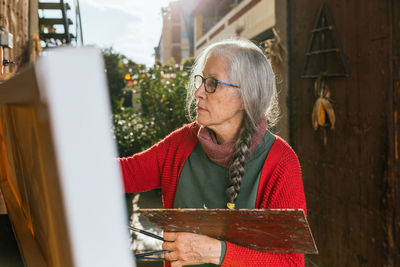 Side view of aged female artists in protective mask painting on canvas in backyard on sunny day in summer