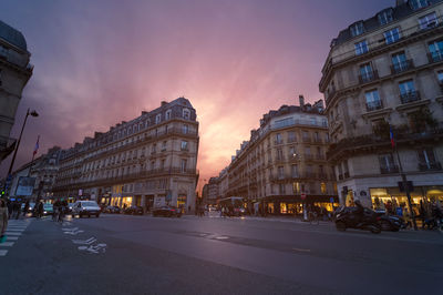 Road by buildings in city against sky during sunset