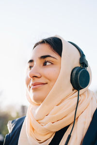 Low angle view of smiling teenage girl listening music through headphones against clear sky