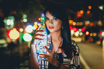 Portrait of young woman holding illuminated bottle while standing outdoors at night