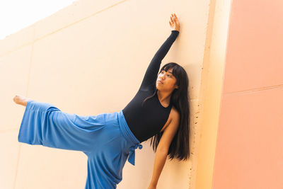 Flexible asian woman with long hair raising leg and stretching while dancing on pavement near orange wall of building on city street
