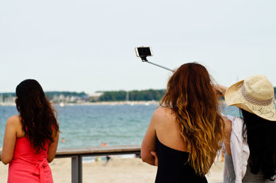 Rear view of friends taking selfie while standing by railing against sea