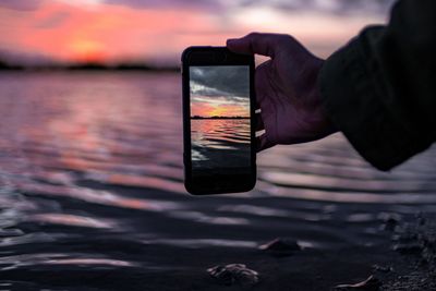 Person holding smart phone by lake against sky during sunset