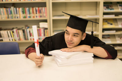 Portrait of student in graduation gown leaning on books at table