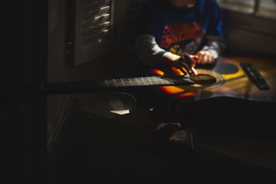 Low section of boy playing guitar while sitting in darkroom at home