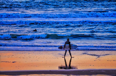 Rear view of man with surfboard standing at beach during sunset