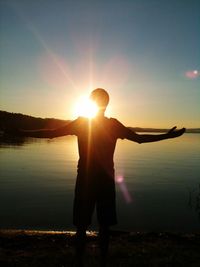 Silhouette man with arms outstretched standing at lakeshore against clear sky