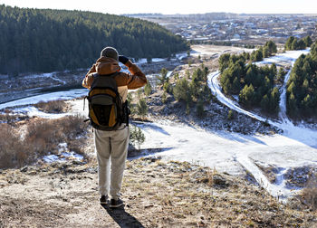 Back view of young man with backpack on mountain looking at view of river and forest winter hiking 