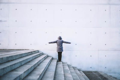 Rear view of woman standing with arms outstretched on steps against gray wall