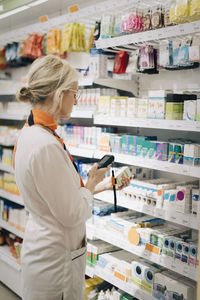 Side view of female owner using bar code reader on medicine standing by rack at store