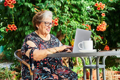 Positive elderly lady in casual outfit and eyeglasses sitting in chair at table while surfing on netbook near cup on saucer and green plants with flowers in yard in daylight