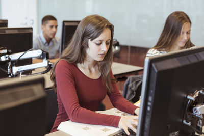 Young female student using computer while sitting with friends in library at high school
