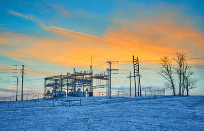 Electricity pylon on snow covered field against sky during sunset