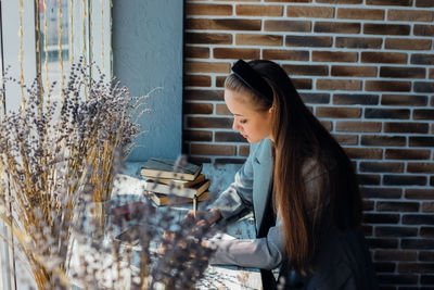 A young woman is sitting at a table by the window and writing in a notebook.