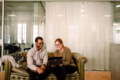 Cheerful business colleagues looking at digital tablet while sitting on sofa in office