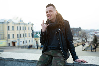 Portrait of smiling young man gesturing horn sign while sitting on retaining wall in city