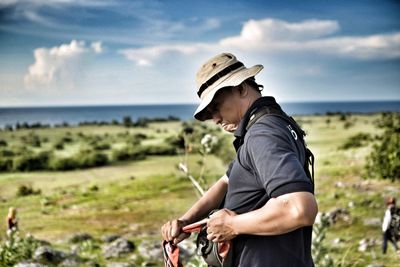 Side view of hiker standing on field by sea against cloudy sky