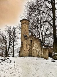 Bare trees on snow covered field by building against sky