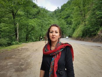 Portrait of beautiful young woman standing at roadside against trees